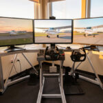 Home flight simulator cockpit deck with yoke, throttle and rudder pedals facing triple monitors at dawn