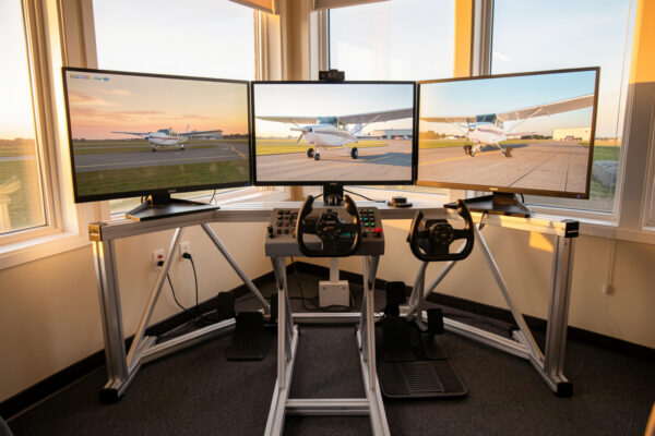Home flight simulator cockpit deck with yoke, throttle and rudder pedals facing triple monitors at dawn